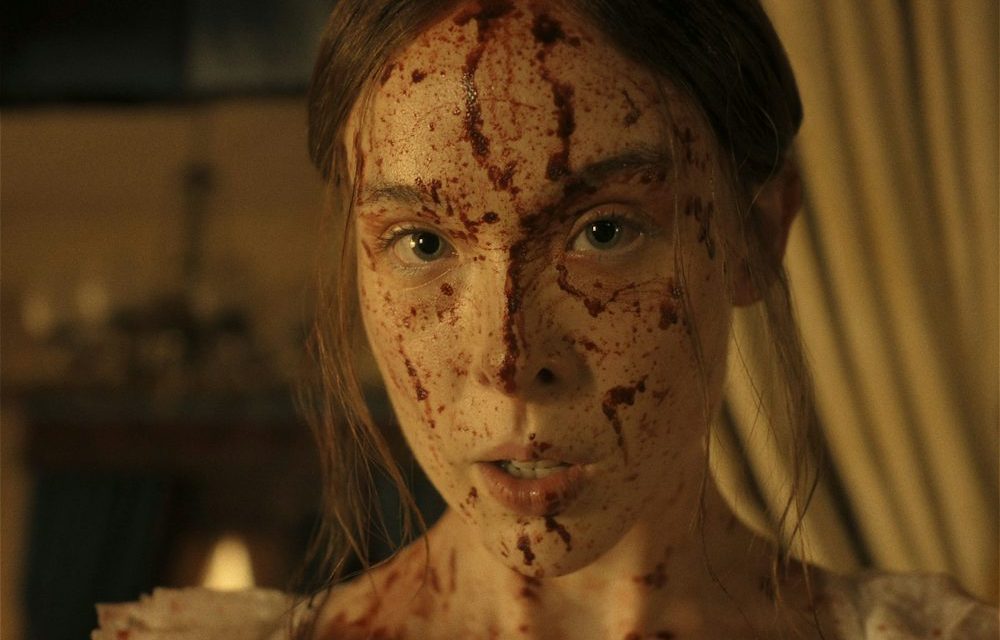 "A young woman with long hair, dressed in light clothing, has her face and upper chest covered in blood, standing indoors with dim warm lighting and blurred curtains and furnishings in the background."