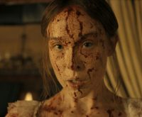 "A young woman with long hair, dressed in light clothing, has her face and upper chest covered in blood, standing indoors with dim warm lighting and blurred curtains and furnishings in the background."
